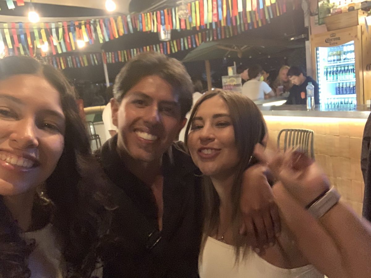 Tres personas riéndose muy juntas en un bar decorado con banderines de colores. Pablo Montes está en el medio.