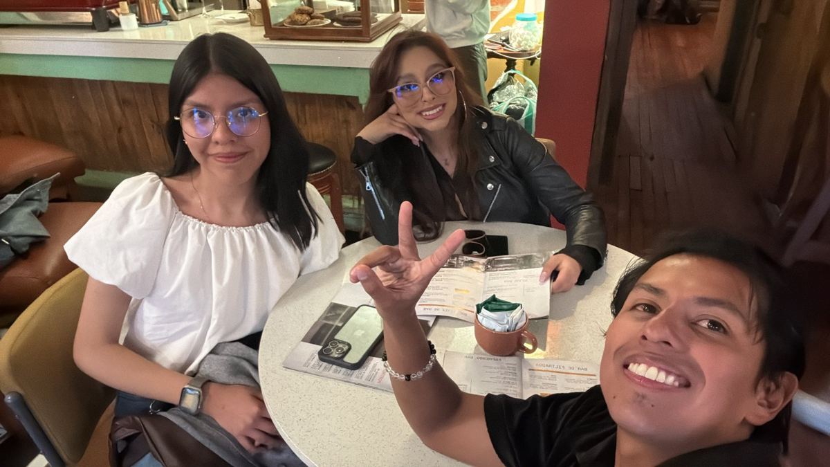 Tres personas sentadas alrededor de una mesa redonda en una cafetería, con las cartas del menú abiertas sobre la mesa. Pablo Montes se toma la foto desde un costado.